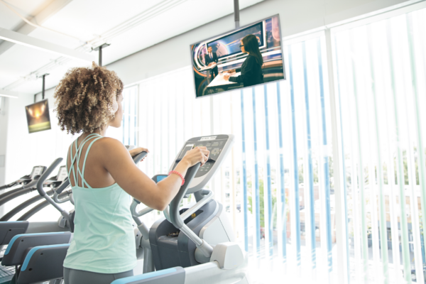 Fit woman on elliptical, engaged with TV entertainment in bright, modern gym setting.